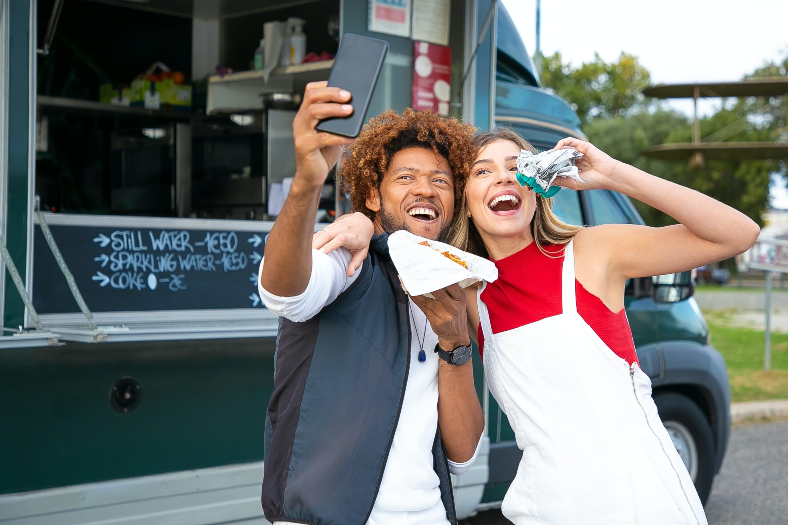 A young couple acting silly and taking a selfie outside of a food truck while they're out on a date.